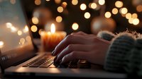 Female hands typing on laptop keyboard in cozy evening atmosphere with glowing candles and bokeh lights creating warm ambiance for remote work or writing.
