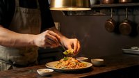 Caucasian male chef in professional kitchen plating pasta dish with garnish, wearing apron. Fine dining food preparation and culinary expertise concept for restaurant, cooking, and gastronomy.
