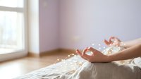 Young woman with medium skin tone relaxing on bed, holding white flower petals in cupped hands, sunlit bedroom with lavender walls, wellness and self-care concept.