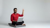 Smiling Middle Eastern businessman in red shirt sitting cross-legged with laptop pointing up, expressing success and achievement in modern workspace.