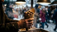 Street food vendor serving roasted chestnuts in paper cone at winter market with warm lighting and blurred crowd background creating cozy atmosphere.