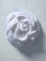 Swirled white cream or foam texture forming rose-like pattern against light background, suitable for cosmetic and beauty product advertising.
