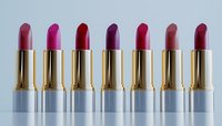 Row of luxurious lipsticks in metallic gold cases displaying various shades of pink, red and purple against light background, makeup cosmetics arrangement.