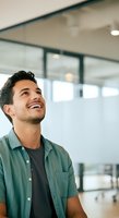 Young man with dark hair and olive complexion, wearing a teal short-sleeved shirt over a dark t-shirt, smiling while looking upward in a modern office environment with glass partitions and natural lighting.