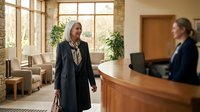 Professional mature businesswoman in elegant coat walking through modern hotel lobby with receptionist at wooden front desk during daytime visit.