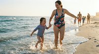 Mother and daughter enjoying summer beach vacation running through ocean waves at sunset with family in background.