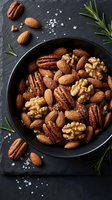 Mixed nuts in black bowl with sea salt and rosemary on dark slate background for healthy snacking and gourmet cooking recipes.