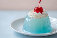 Blue jelly dessert topped with whipped cream and a red cherry on a white plate, soft pastel background, sweet treat for food blogs and menus.