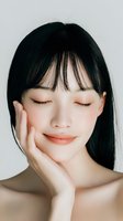 Young Asian woman with closed eyes and gentle smile touching face against white background. Portrays serenity and natural beauty for skincare advertising.