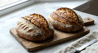 Two freshly baked artisan sourdough bread loaves with scored crusts on wooden cutting board near window, rustic bakery style for food photography and cooking.