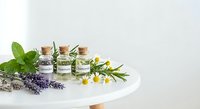 Essential oils in glass bottles with cork stoppers surrounded by fresh herbs including mint, lavender, and chamomile on white surface for aromatherapy.