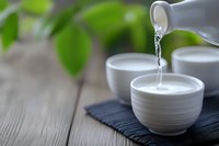 Pouring clear liquid from ceramic pitcher into white bowl on dark slate with green foliage background creating peaceful zen atmosphere for wellness content.