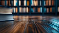 Stack of books on wooden table against blurred library bookshelf background with warm lighting and shallow depth of field creating cozy academic atmosphere.