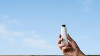 Hand holding sunscreen bottle against clear blue sky and horizon, ideal for skincare, sun protection, and summer wellness marketing campaigns.