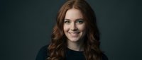 Professional young Caucasian businesswoman with curly red hair smiling confidently in dark sweater against gray background for corporate headshots.