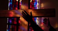Hand reaching toward wooden cross with colorful stained glass window light creating spiritual atmosphere for worship and prayer concepts.