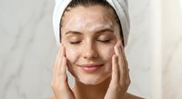 Young woman applying facial cleanser with closed eyes and white headband in bathroom skincare routine for healthy glowing skin and daily beauty care.