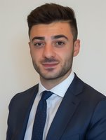 Young Mediterranean businessman in navy suit and polka dot tie smiling confidently against light background, professional corporate headshot style portrait.