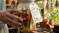 Man holding glass jar with homemade garden infused water, fresh herbs and vegetables inside, labeled tag visible, greenhouse setting with potted plants background.