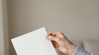 Hand holding blank white paper sheet against neutral wall background for mockup and branding purposes.