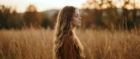 Young woman with long wavy hair in golden wheat field at sunset, peaceful moment in nature, warm autumn light creating dreamy atmosphere for lifestyle content.