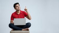 Young Asian man in red polo shirt sitting cross-legged with laptop pointing up having creative idea isolated on white background for design projects.