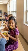 Young African American girl enjoying healthy lunch in school cafeteria, smiling while eating nutritious meal at wooden table with classmates in background.