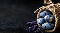 Blue speckled eggs spilling from rustic burlap sack with dried lavender sprigs on dark textured surface for Easter spring decoration and organic farm concepts.