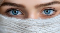Close up view of bright blue eyes with long eyelashes peering over white knitted scarf or face mask, expressing mystery and intrigue in healthcare or fashion context.