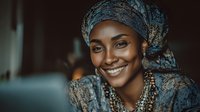 African woman wearing traditional blue headwrap and jewelry smiling warmly while using laptop computer for remote work or online business communication.