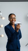 Happy African American businesswoman in navy suit holding paper airplane in modern office with creative lighting and minimalist decor.