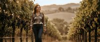 Young woman walking through vineyard rows during golden hour sunset with rolling hills in background for wine tourism and agricultural lifestyle content.