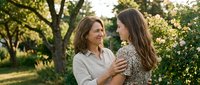 Two happy women embracing outdoors in sunny park setting with green trees background, showing genuine friendship and emotional connection between generations.