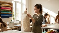 Young Caucasian female fashion designer adjusting garment on dress form in bright modern atelier workspace with fabric samples and sewing equipment.