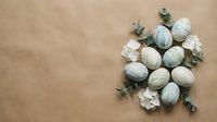 Easter eggs decorated in soft green and white tones with eucalyptus branches and flowers on beige background for spring holiday celebration.