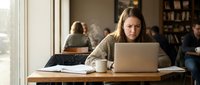 Young woman working on laptop in cozy cafe with warm lighting. Professional freelancer or student studying in coffee shop environment with books and cup.