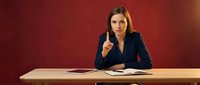 Confident young Caucasian businesswoman in navy suit sitting at desk, raising finger to make a point, red background, serious expression.
