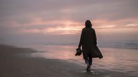 Woman in dark coat walking into ocean at sunset, silhouetted against golden sky and calm water, contemplative mood for editorial and lifestyle use.