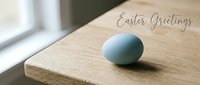 Soft blue Easter egg on a wooden table near a window with Easter Greetings text, minimalist spring holiday card design.