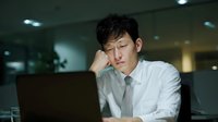 Young Asian businessman looking tired and stressed while working late at office computer, resting head on hand in dimly lit corporate environment.