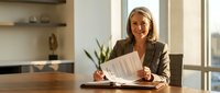 Confident senior Caucasian businesswoman signing documents at office desk, smiling, holding papers in professional meeting room setting.