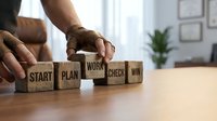 Business professional arranging wooden blocks spelling START PLAN WORK CHECK WIN on desk, representing strategic planning and success methodology for corporate growth.