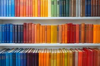 Colorful books arranged by spectrum colors on white library shelves, from blue through orange and red creating rainbow effect. Academic collection background.