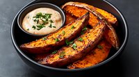 Roasted sweet potato wedges seasoned with herbs and spices served in black ceramic bowl with creamy dipping sauce garnished with fresh parsley on dark background.