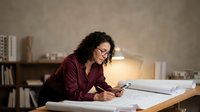 Professional woman architect with curly hair working late on blueprints at drafting table in modern office with warm lighting for design projects.