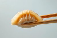 Asian dumpling held with wooden chopsticks against light background, showing translucent wrapper and pleated edge detail in close-up view.