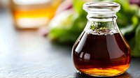 Dark amber balsamic vinegar in glass bottle with fresh herbs, blurred salad greens and oil in background for gourmet cooking and food styling.