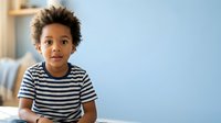 Young boy with curly brown hair and medium skin tone wearing striped shirt, looking at camera with neutral expression against blue background for portrait use.
