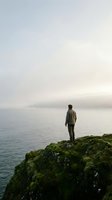 Man standing on moss-covered rocky cliff overlooking calm misty lake, wearing casual jacket, contemplating serene water landscape with distant mountains shrouded in fog.