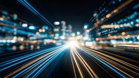 Dynamic city street at night with motion blur light trails from traffic creating streaks of blue and orange illumination through urban canyon of buildings.